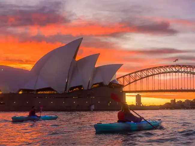 Kayakers paddling past Sydney Opera House during vivid orange sunset on the harbour.
