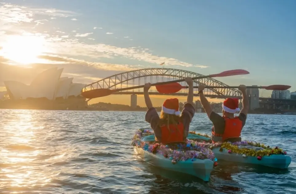kayakers in santa hats enjoy a guided kayaking tour on sydney harbour passing the sydney opera house.